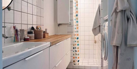 Moderne badeværelse med egetræsbordplade og vask, vaskesøjle med vaskemaskine og tørretumbler. Hvidt flisebelægning med farverig bruseforhæng i polka dots. Fokus på effektiv opbevaring og funktionalitet i et lille badeværelse.