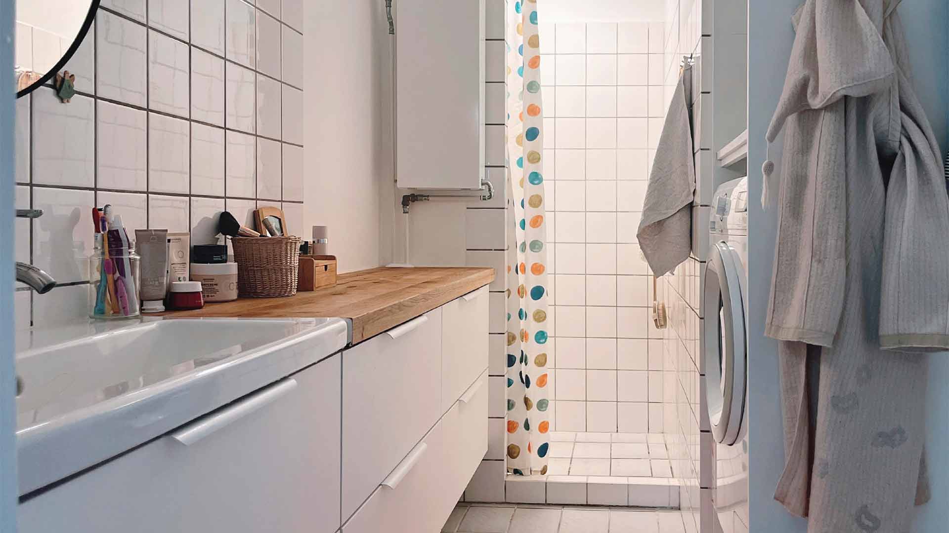 Moderne badeværelse med egetræsbordplade og vask, vaskesøjle med vaskemaskine og tørretumbler. Hvidt flisebelægning med farverig bruseforhæng i polka dots. Fokus på effektiv opbevaring og funktionalitet i et lille badeværelse.